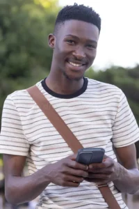 african-american-man-smiling-holding-phone-park-wearing-striped-tee-crossbody-strap-portrait-outdoor-nature-device-casual-461716142